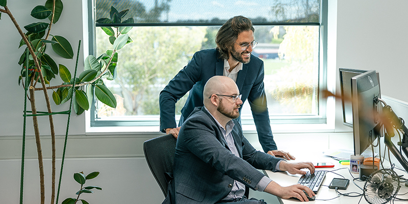 Two male IP professionals in suits sit at an office desk use a computer by a window