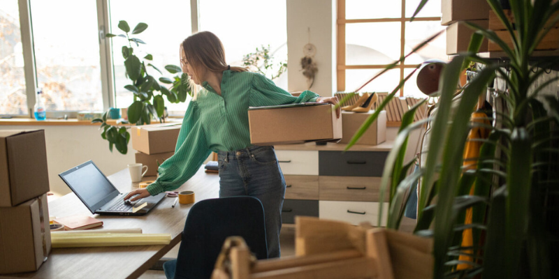 Woman juggles a box and working on her laptop in a studio surrounded by more boxes and plants
