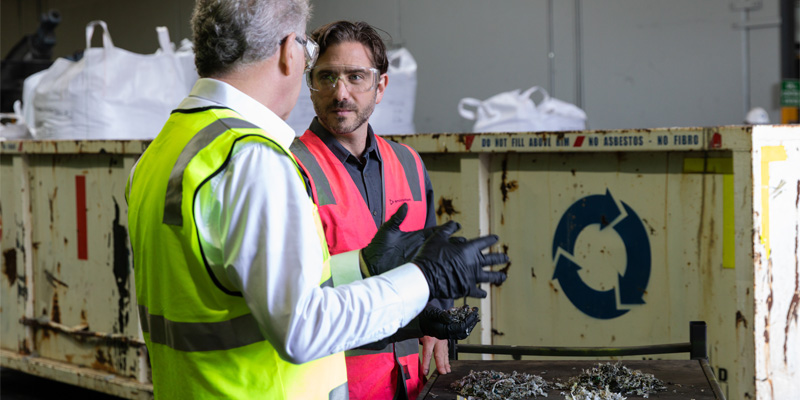 Two people wearing high-visibility safety vests and protective gloves discussing materials at a recycling facility.