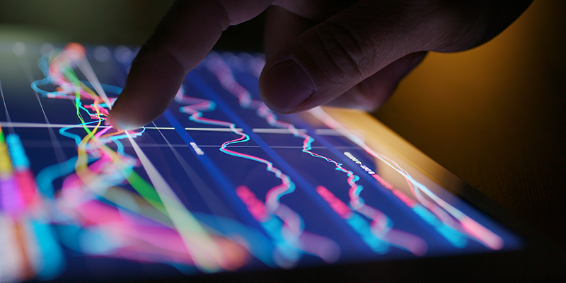 A person uses their finger to interact with a tablet displaying colourful financial data charts and trend lines on a dark screen.