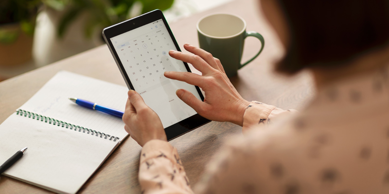 A woman's hands scrolling a calendar on a tablet device. A notebook and coffee cup are in the background.